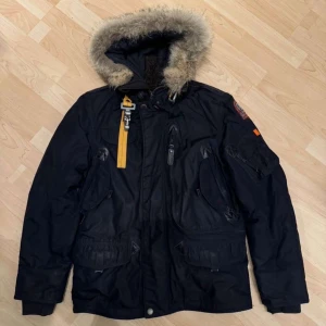Svart parkajacka från Parajumpers - Säljer en svart parkajacka från Parajumpers med pälsfodrad huva och flera praktiska fickor. Jackan har en gul detalj vid dragkedjan och en broderad patch på ärmen. Perfekt för kalla dagar.