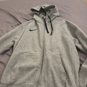 Grå hoodie Nike - Säljer en grå hoodie från Nike med dragkedja och svart logotyp på bröstet. Tröjan har en justerbar huva med svarta snören och praktiska fickor framtill. Perfekt för en avslappnad stil.