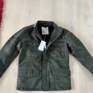 Camouflage Zadig Voltaire jacka - Säljer en grön camouflagejacka med dragkedja och knappar. Jackan har flera praktiska fickor och är långärmad. Perfekt för en cool och avslappnad stil.