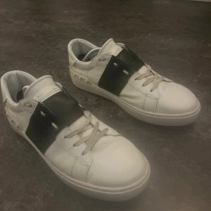 Valentino open  - Snygga vita sneakers med en bred svart rem över snörningen från Valentino. Skorna har dekorativa nitar runt hälen och en klassisk låg profil. Perfekta för en stilren look. Valentino open storlek 43 inget og följs med 