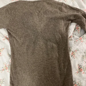 Brun stickad klänning med strassdetaljer - Jättemysig brun/grå typ stickad klänning med v-ringning och långa ärmar. Baksidan pryds av söta strassdetaljer i formen av ett par änglavingar på ryggen. Perfekt för kyligare dagar när du vill vara både bekväm och fin. Klänningen är i bra skick och jag har nästan aldrig använt den🩷Den är i S/M men är ganska liten i storleken, passar mig som har typ XS. Hör av er om ni har några frågor🫶🏼