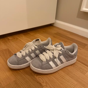 Grå Adidas Campus sneakers - Snygga grå sneakers från Adidas med klassiska vita ränder på sidan. Skorna har en stilren design med snörning och är tillverkade i mocka. Perfekta för en avslappnad stil.