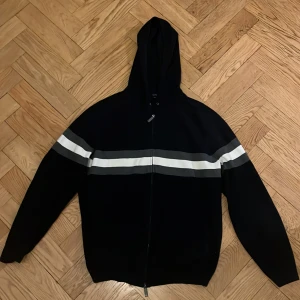 Mörk blå hoodie med ränder - Säljer en stilren blå hoodie med dragkedja och huva. Den har vita och grå ränder över bröstet och ärmarna. Perfekt för en avslappnad stil.