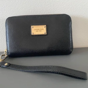 Svart plånbok från Michael Kors - Snygg svart plånbok från Michael Kors med guldfärgad logoplatta och dragkedja. Tillverkad i skinn med en praktisk handledsrem. Perfekt för att hålla ordning på dina kort och kontanter med stil. Svart plånbok med avtagbart handtag från Michael Kors.  Plånboken är använd och metallmärkena är lite slitna i övrigt i fint begagnat skick.  Mått Längd: ca 14 cm Bredd: ca 9 cm  Kommer från ett djur och rökfritt hem.