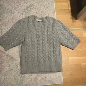 Grå stickad tröja - Säljer en snygg grå stickad tröja från Zara med flätmönster och pärldetaljer. Tröjan har trekvartsärmar och ribbade kanter. Den är i storlek S helt ny 