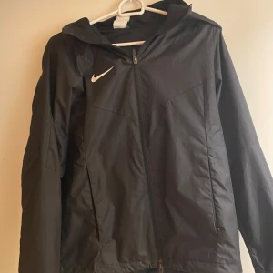 Svart vindjacka från Nike - Snygg svart vindjacka från Nike med dragkedja och huva. Perfekt för blåsiga dagar. Jackan har en stilren design med en liten Nike-logga på bröstet. Passar bra för både träning och vardag.