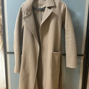 Kappa  - Snygg beige kappa från new torksr  med klassisk krage och långa ärmar. Den har en stilren design med två stora fickor framtill. Perfekt för höst och vår. 