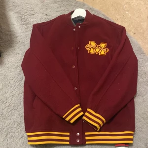 Vinröd bomberjacka från H&M - Snygg vinröd bomberjacka från H&M med gula detaljer och tryck på bröstet. Jackan har knappar framtill och ribbade muddar i gult och rött. Perfekt för en sportig look! Använt minst 3 gånger och sen dess har den bara varit i min garderob. 