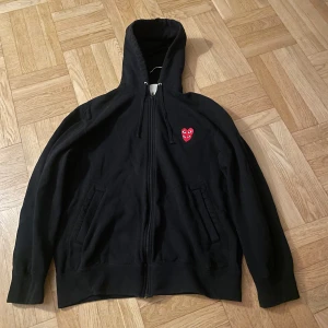Svart hoodie från Comme des Garçons - Säljer en svart hoodie från Comme des Garçons med dragkedja och det ikoniska röda hjärtat på bröstet. Perfekt för en stilren och bekväm look. Huvan har snörning och tröjan är tillverkad i bomull.