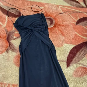 Mörkblå klänning med smala axelband - Elegant mörkblå klänning med smala axelband och draperad detalj framtill. Perfekt för en stilren look. Klänningen har en figurnära passform och är tillverkad i ett mjukt material som ger en fin siluett.