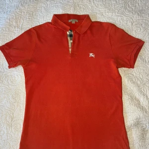 Röd pikétröja från Burberry - Snygg röd pikétröja från Burberry med klassisk krage och knappslå med rutigt mönster. Tröjan är i mycket bra skick bara använda några gånger, Perfekt för en stilren look.