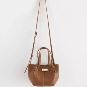 Brun väska - Snygg brun axelväska i skinn med justerbar axelrem och praktiska handtag. Väskan har en elegant gulddetalj framtill och knytband på sidorna. Perfekt för att bära dina nödvändigheter med stil. Väskan är slutsåld på hemsidan! Väskan är helt oanvänd och prislappen sitter fortfarande på! 