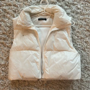 Vit dunväst från Shein - Säljer en snygg vit dunväst från Shein med dragkedja framtill. Västen har en hög krage och justerbar dragsko i nederkant för en perfekt passform. Ett brännmärke på ena sidan av västen.