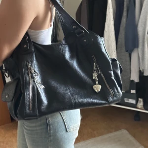 Svart axelväska med detaljer - Snygg svart axelväska i skinn med dragkedjor och dekorativa hängen. Väskan har flera fack och en praktisk storlek för vardagsbruk. Perfekt för att hålla ordning på dina saker med stil.