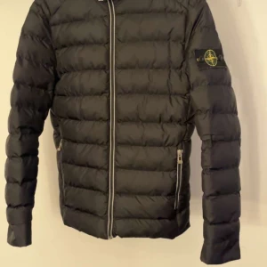 Svart pufferjacka från Stone Island - Snygg svart pufferjacka från Stone Island med dragkedja framtill och deras ikoniska märke på ärmen.  Jackan har en quiltad design och långa ärmar, perfekt för kyligare dagar. Den är både stilren och funktionell. Byten går också, Skick 10/10
