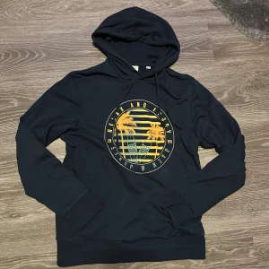 Svart hoodie från Jack & Jones - Säljer en svart hoodie från Jack & Jones med storlek S och ett coolt tryck av en solnedgång och palmer på framsidan. Tröjan har en klassisk huva med dragsko och långa ärmar. Perfekt för en avslappnad stil.