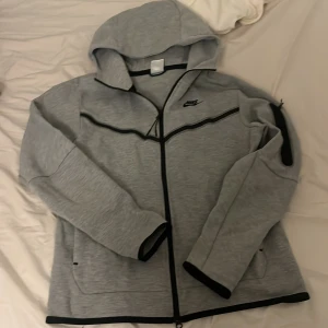 Nike tech fleece  - Säljer en grå hoodie från Nike med dragkedja och huva. Den har svarta detaljer och en liten Nike-logga på bröstet. Perfekt för en avslappnad stil.