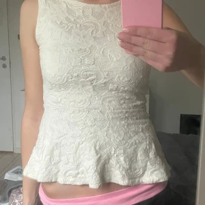 Vit spetslinne - Säljer ett vitt spetslinne med blommigt mönster och volangdetalj i nederkant. Linnet är ärmlöst och har en elegant passform som framhäver figuren. Perfekt för en stilren look.