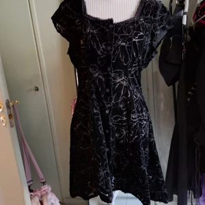 Svart sammet klänning med blommönster - Snygg svart klänning i sammet med ett subtilt blommönster. Klänningen har korta ärmar och en rundad halsringning med dekorativ kant. Perfekt för en elegant look.