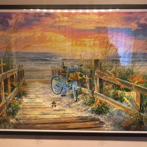 Pussel med strandmotiv - Ett vackert pussel med ett strandmotiv där en blå cykel står parkerad på en träbrygga vid solnedgången. Omgiven av blommor och gräs, med havet i bakgrunden. Perfekt för den som älskar somriga och lugna landskap.