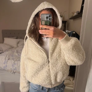 Vit teddyjacka med huva - Mysig vit teddyjacka med huva och dragkedja framtill. Perfekt för kyliga dagar, den har en fluffig textur och ribbade muddar vid ärmslut och nederkant. Jackan är både stilren och bekväm, idealisk för en avslappnad look.