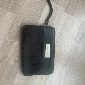 Svart stor plånbok från Calvin Klein - Snygg svart plånbok från Calvin Klein med ett diskret CK-mönster. Den har en praktisk dragkedja och flera fack för kort och kontanter. Perfekt för att hålla ordning på dina värdesaker med stil. Sminkfläck invändigt som inte går bort samt att dragkedjan invändigt börjar släppa på färg. Calvin Klein märket framtill har små repor men märks ej av. Ca 14x22 cm.