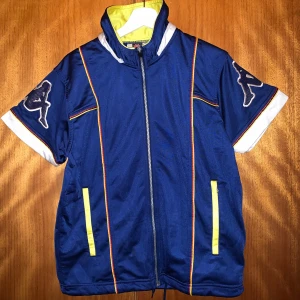 Blå kortärmad jacka från Kappa - Säljer en retro blå kortärmad väst från Kappa med dragkedja och vita detaljer. Jackan har gula och orangea linjer samt Kappas logga på ärmarna. Perfekt för en sportig look!