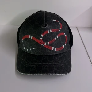 Svart keps med ormmönster - Snygg svart keps med ett iögonfallande ormmönster i rött och vitt. Kepsen har ett diskret mönster i tyget och justerbar rem baktill. Perfekt för en cool och avslappnad stil.