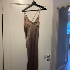 Brun långklänning med smala axelband - Elegant brun långklänning med smala axelband och v-ringning. Klänningen har en knytning i ryggen och är tillverkad i ett glansigt material som ger en lyxig känsla.