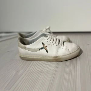 Vita sneakers från Axel Arigato - Snygga vita sneakers från Axel Arigato med broderad fågel på sidan. Skorna har en klassisk design med snörning och är tillverkade i skinn. Perfekta för en stilren look.