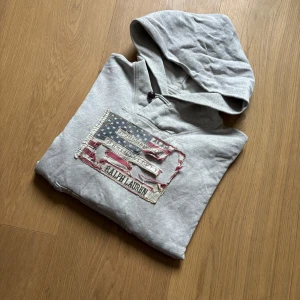Grå hoodie från Ralph Lauren - Tja! Säljer en schysst Ralph Lauren hoodie med sliten flagga som ger en unik vibe. Storlek M kan nog passa en S. Finns tyvärr en liten defekt på bild 4, men annars i fint skick! Hör av dig vid frågor eller liknande✌️
