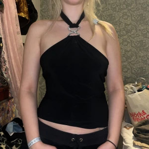 Svart halterneck topp - Snygg svart halterneck topp med dekorativt spänne framtill. Perfekt för en stilren look. Toppen är ärmlös och har en tight passform.