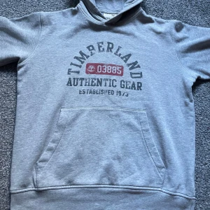 Grå hoodie från Timberland - Säljer en grå hoodie från Timberland med tryck på framsidan. Tröjan har en klassisk känguruficka och en bekväm huva. Passar både storlek M och S.  