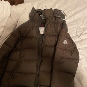Khaki dunjacka från Moncler - Säljer en snygg svart dunjacka från Moncler. Jackan har en dragkedja framtill och en broderad logotyp på ärmen. Perfekt för kyliga dagar med sin quiltade design som håller värmen. Den är stilren och passar bra till olika outfits.