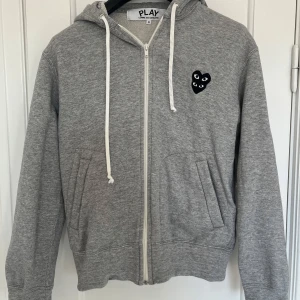 Grå hoodie från Comme des Garçons Play - Säljer en stilren grå hoodie från Comme des Garçons med dragkedja och huva. Den har ett ikoniskt hjärtmärke med ögon på bröstet. Perfekt för en casual look.