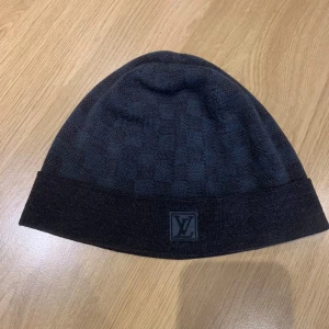 Mörkblå mössa från Louis Vuitton - Snygg mörkblå mössa från Louis Vuitton med diskret rutigt mönster och uppvikt kant. Perfekt för att hålla värmen med stil. Märkeslogga framtill ger en exklusiv touch. Dessutom så är den som ny.