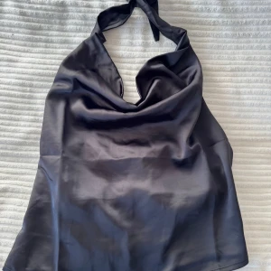 Svart halterneck topp från Weekday - Snygg svart halterneck topp från Weekday i ett glansigt material. Perfekt för en stilren look med en elegant touch. Toppen har en draperad framsida och knytning i nacken.