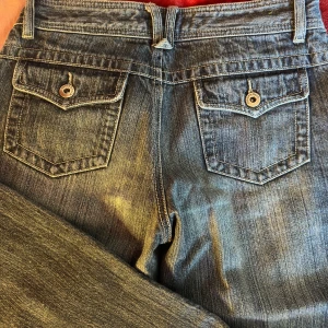 Snygga långa lågmidjade jeans - Snygga blå jeans från Track & Field med knappar på bakfickorna. De har en rak passform och lågmidjade. Mått innerbenslängd 85cm och midjan 37cm. Inte så stretchiga. Säljer då dem är för små för mig så inga bilder på.
