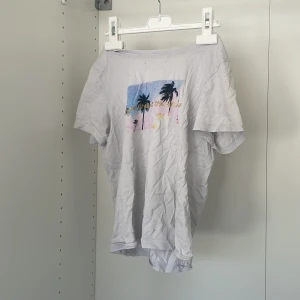 Vit t-shirt med tryck från Firefly - Säljer en vit t-shirt från Firefly med ett färgglatt tryck av palmer och text på framsidan. T-shirten har korta ärmar och är perfekt för en avslappnad stil. Materialet är mjukt och bekvämt.