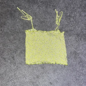 Gul blommig linnetopp från Stories - Söt gul linnetopp från Stories med små blommor i vitt och svart. Toppen har smala axelband som kan knytas och en smockad design för en bekväm passform. Perfekt för varma sommardagar!