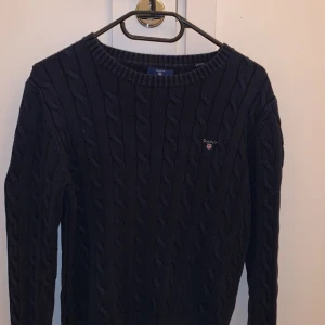 Mörkblå stickad tröja från GANT - Säljer en snygg mörkblå stickad tröja från GANT med kabelstickat mönster. Tröjan är lite sliten i halsen men är annars i mycket bra skick.