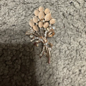 Elegant brosch i form av en blombukett - Säljer en elegant brosch i form av en blombukett med ljusrosa stenar och gnistrande detaljer. Broschen har en vacker rosett och är perfekt för att ge en extra touch till din outfit.