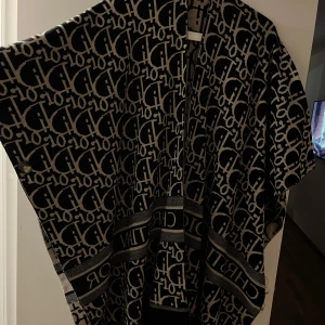 Svart och beige poncho  - Snygg poncho i svart och beige med ett iögonfallande mönster av märkets logotyp. Ponchon har en lös passform och är perfekt för att ge en lyxig touch till din outfit. Den är tillverkad i ett mjukt material och har knappar på sidorna för extra stil och funktionalitet. Använd 1 gång. Går även att ha på båda sidorna om man vill ha annan färg. 