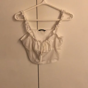 Vit croppad topp från Shein - Säljer en söt vit croppad topp från Shein med volangdetaljer på axelbanden och en knytning framtill. Perfekt för varma sommardagar eller som ett lager under en jacka. Toppen är i storlek M men skulle säga att den passar XS-L🥰