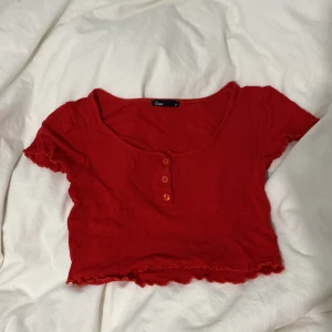 Röd ribbad topp från Elam - Säljer en snygg röd ribbad topp från Elam. Den har korta ärmar och tre knappar framtill. Toppen är croppad med en vågig kant som ger en extra touch. Perfekt för en trendig look!