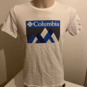 Vit t-shirt från Columbia - Snygg vit t-shirt från Columbia med ett stort blått och svart grafiskt tryck på framsidan. T-shirten har korta ärmar och en rund halsringning. Perfekt för en avslappnad stil.