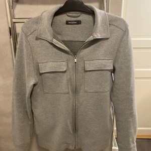 Grå overshirt från dressmann - Snygg grå overshirt från dressmann med dragkedja och två stora fickor framtill. Jackan har en klassisk krage och långa ärmar, perfekt för en stilren look. Passar bra till både vardag och mer uppklädda tillfällen. 