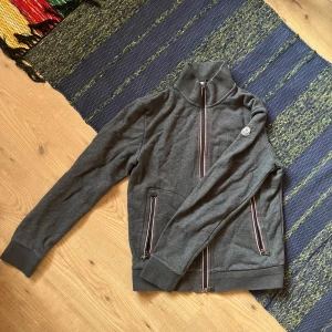 Moncler Zip Tröja - Säljer en stilren grå jacka från Moncler med dragkedja och ribbade muddar. Jackan har ett broderat Moncler-märke på ärmen och detaljer i rött och vitt längs dragkedjan. Storlek Large men passar S/M,  knappt använd! Kan gå ner i pris vid snabb affär!