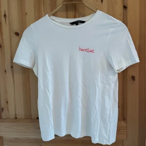 Vit t-shirt från Vero Moda - Säljer en stilren vit t-shirt från Vero Moda med texten 'heartbeat' broderad i rött på bröstet. T-shirten har en klassisk rund hals och korta ärmar. Perfekt för en avslappnad look.