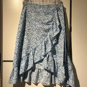 Blommig blå omlottkjol - Charmig blå omlottkjol med blommigt mönster och volangdetaljer. Kjolen har en knytning i midjan som ger en fin passform. Perfekt för en somrig look!